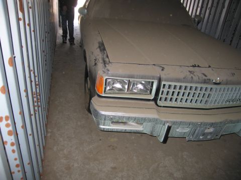 1991 Ford LTD Crown Victoria - Photo of the dust-covered front of a tan Chevrolet sedan parked in a narrow storage container.