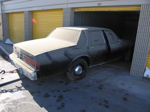 1991 Ford LTD Crown Victoria - Photo of the rear three-quarter view of a dusty black Chevrolet sedan outside yellow storage units.