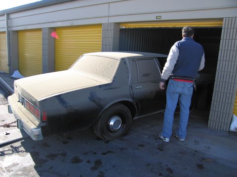 1991 Ford LTD Crown Victoria - Photo of a person opening the door of a dusty black Chevrolet sedan outside storage units.