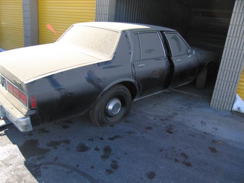 1991 Ford LTD Crown Victoria - Photo of the rear side of a dust-topped black Chevrolet sedan outside storage garage units.
