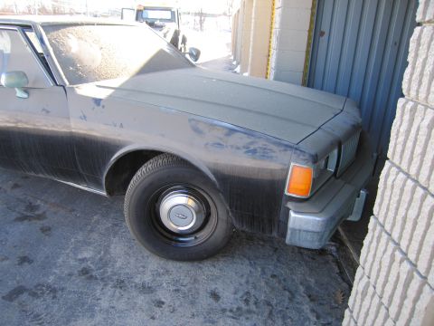 1991 Ford LTD Crown Victoria - Photo of the front fender and headlight of a dusty black Chevrolet sedan beside a garage.