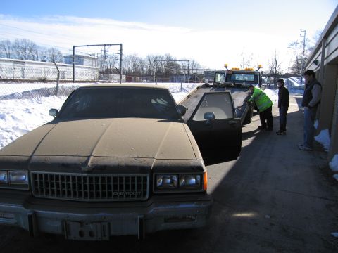 1991 Ford LTD Crown Victoria - Photo of a dust-covered Chevrolet sedan loaded beside a flatbed tow truck as people watch in snow.