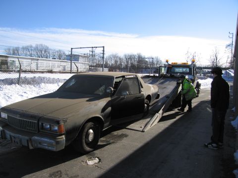 1991 Ford LTD Crown Victoria - Photograph of a workers loading a dirty Chevrolet Caprice onto an inclined flatbed tow truck.