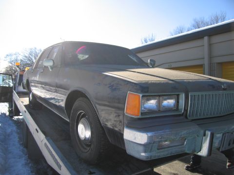 1991 Ford LTD Crown Victoria - Photograph of a grimy Chevrolet Caprice sedan loaded on a flatbed tow truck beside storage units.