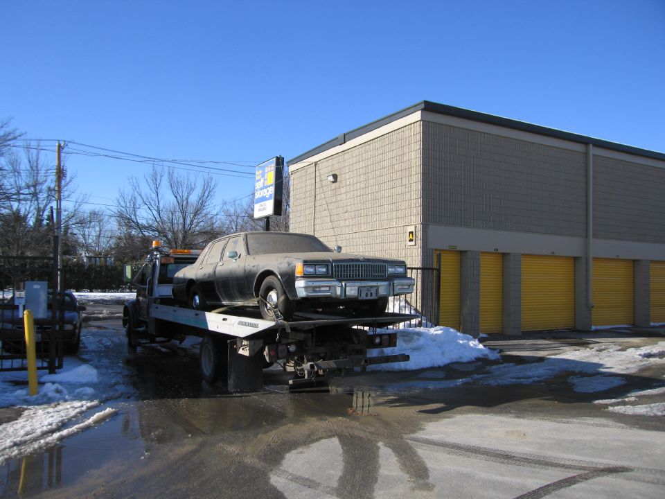 Dusty sedan on a flatbed tow truck near a storage facility
