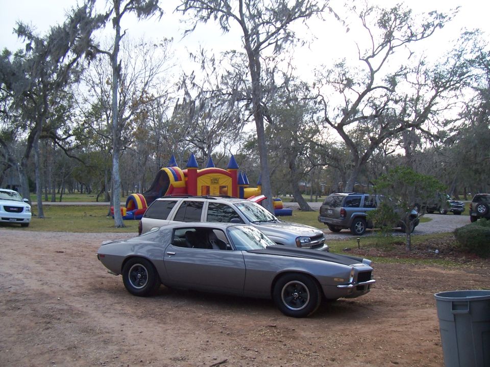 Gray 1970s Chevrolet Camaro parked on dirt lot with bouncy castle and SUVs in background
