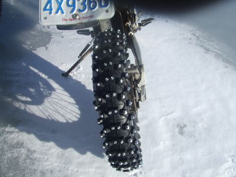 1995 Ford Mustang - A motorcycle tire equipped with studs is shown on a snow-covered ground, with a license plate in the background.