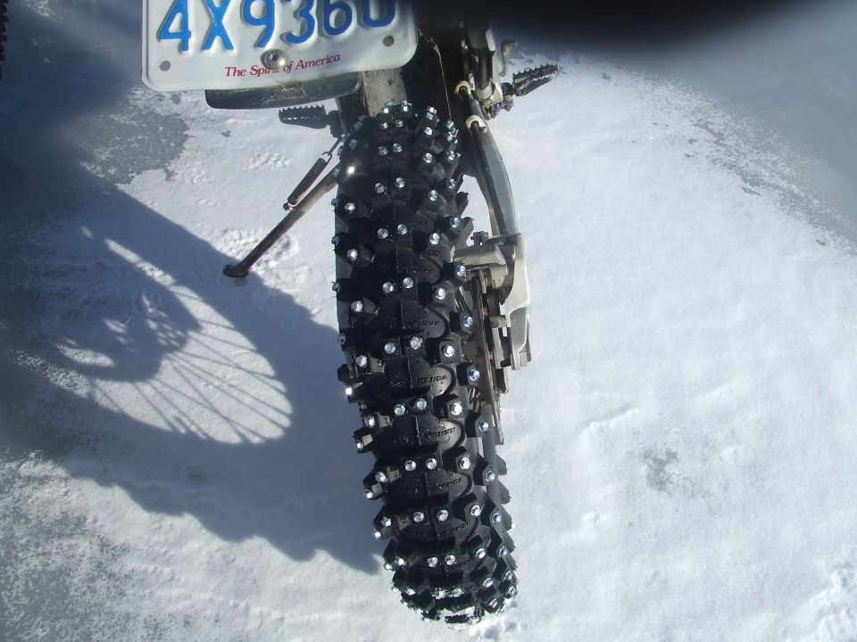 Rear tire of a motorcycle with studs for snow, parked on a snowy surface. License plate visible