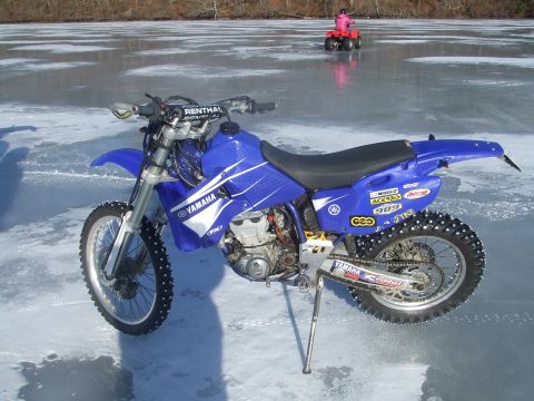1995 Ford Mustang - A blue Yamaha dirt bike equipped with studded tires sits on a frozen surface, with an ATV visible in the distance.