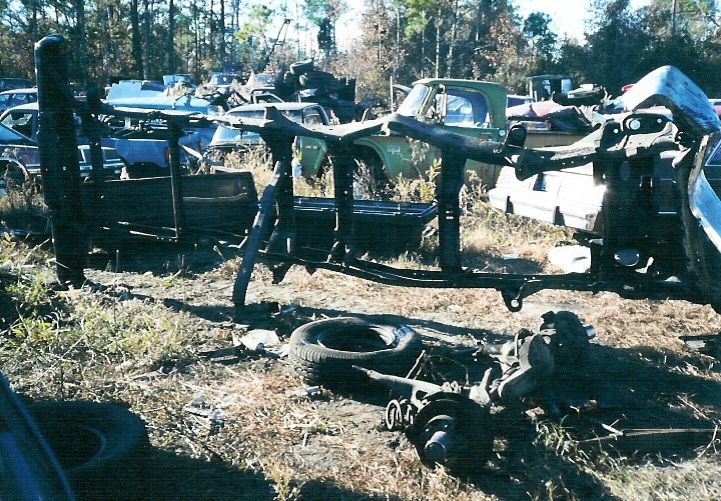 Stripped truck frame and front suspension parts lying in a wooded salvage yard