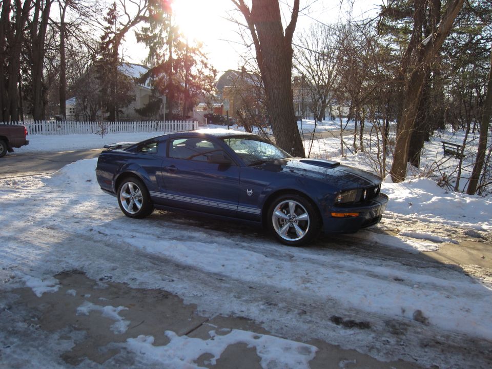 Blue Ford Mustang parked on snowy driveway, sun glare through trees