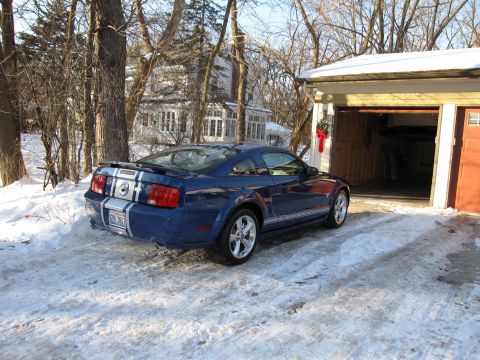 1992 Ford Bronco - A blue Ford Mustang with white racing stripes sits in a snowy driveway next to an open garage.