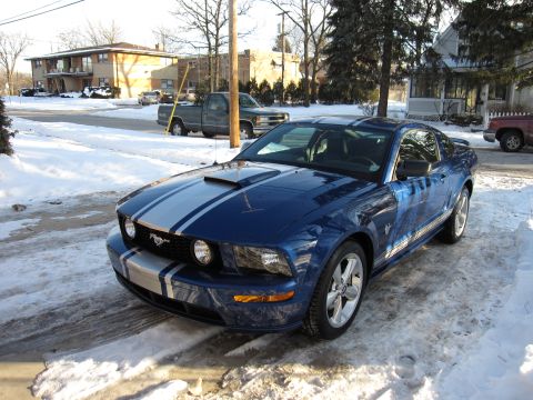 1992 Ford Bronco - A blue Ford Mustang with racing stripes sits on a snow-covered road in front of apartment buildings.