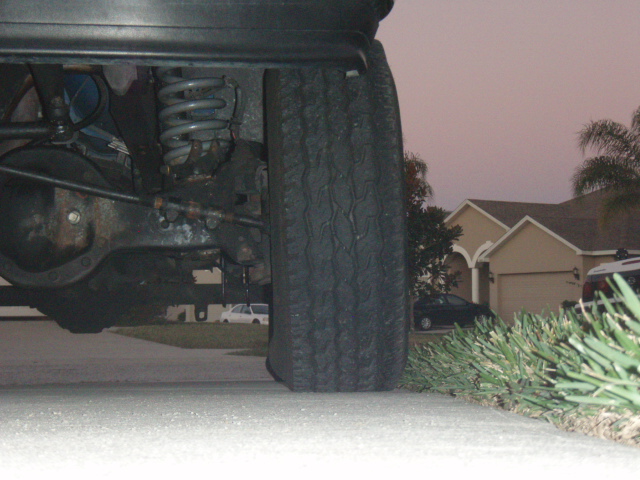 Undercarriage view of a vehicle's rear axle, suspension, and tire. Houses and cars are visible in the background