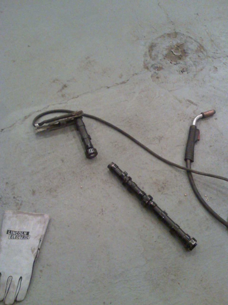Camshaft and welding torch on concrete floor next to a Lincoln Electric glove