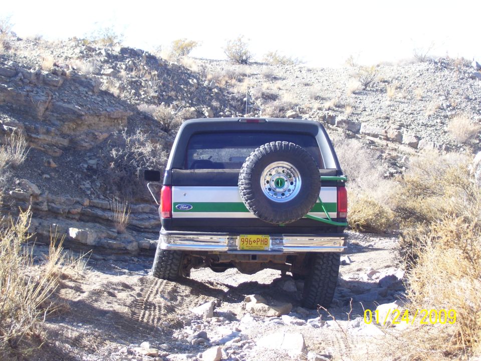 Rear view of a black and silver Ford Bronco with green stripes, spare tire mounted on the back, driving on a rocky trail