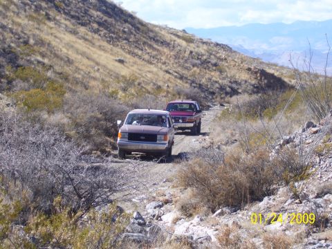 1993 Ford Bronco - Two Ford trucks navigate a dusty trail through a barren, mountainous terrain.