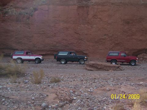 1993 Ford Bronco - Three Ford Broncos drive along a dirt road in a desert landscape with a large red rock formation.