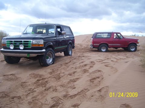 1993 Ford Bronco - Two Ford Broncos are parked on a sandy dune under a cloudy sky. The black Bronco is in the foreground.