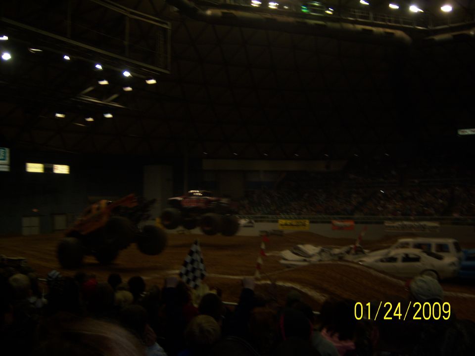 Two monster trucks in mid-air at an indoor event with a crowd watching