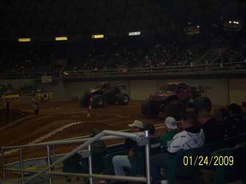 1996 Ford Bronco - Monster trucks are prepared for an event in an indoor arena, with a crowd watching from the stands.