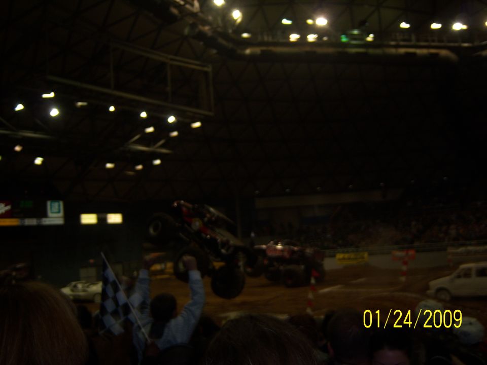 Monster truck airborne over dirt arena, with another truck behind it and spectators in foreground