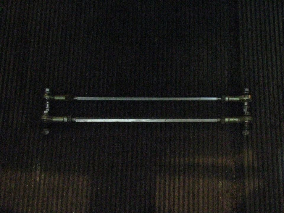 Two metal tie rods with threaded ends and ball joints, laid horizontally against a dark, textured surface