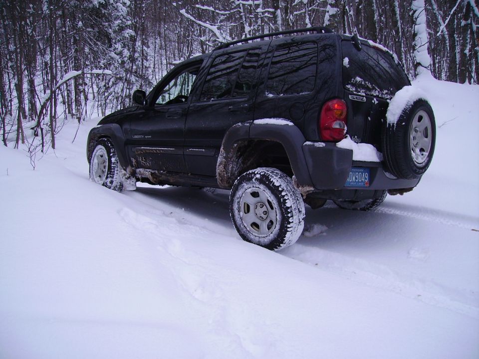 Black Jeep Liberty with spare tire on rear, driving in snow. License plate reads DV9049