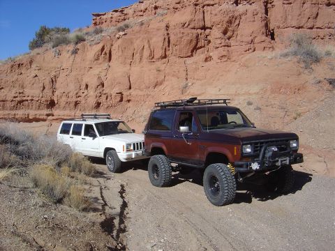 1996 Ford F-250 - Two off-road vehicles navigate a dusty trail through a canyon with steep red rock formations.