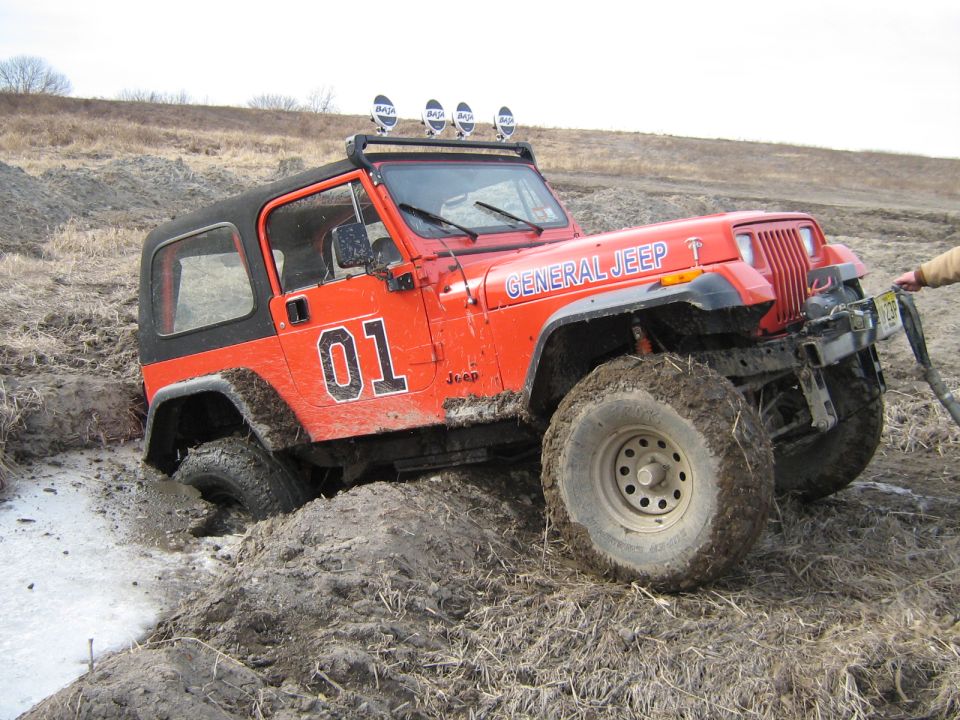 Orange Jeep Wrangler with 'General Jeep' and '01' on the side is stuck in mud, with a person pulling on a winch line