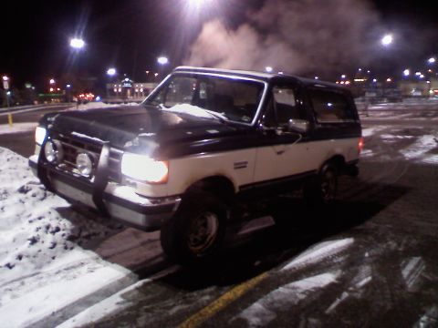 1988 Ford Bronco - This two-tone Ford Bronco is parked in a snowy lot at night, with exhaust fumes rising.