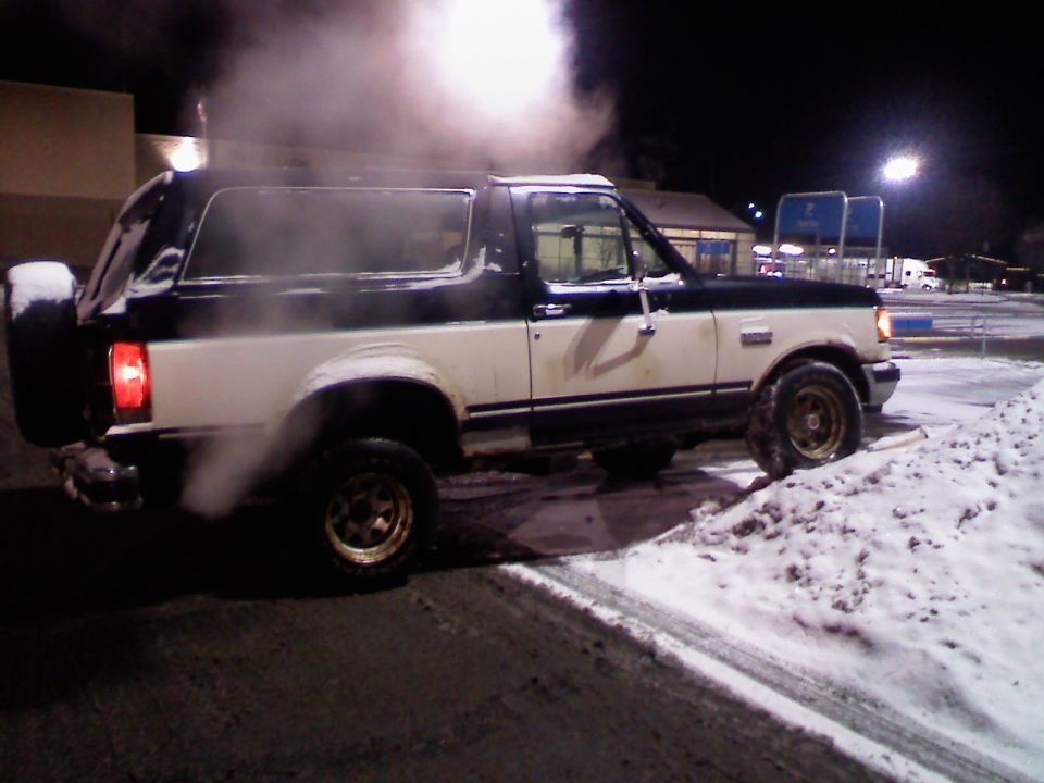 Two-tone white and black Ford Bronco with a spare tire on the back, emitting smoke in a snowy parking lot