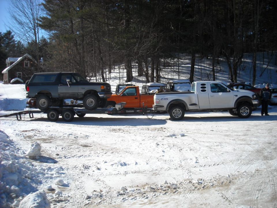 Gray Ford Bronco on trailer being towed by orange truck, white pickup truck behind it