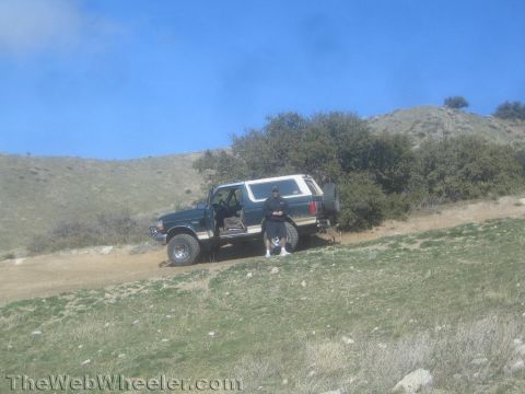 1990 Ford Bronco - dark full-size SUV with white top parked on a dirt trail on a green hillside under blue sky, a person standing at its open door.