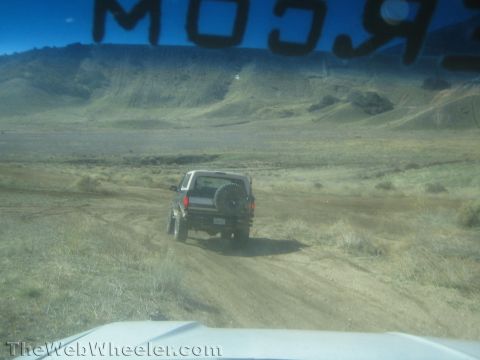 1990 Ford Bronco - View through a windshield of an SUV with a rear-mounted spare tire driving away on a dirt road across a dry desert basin.