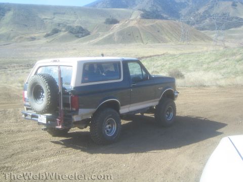 1990 Ford Bronco - lifted dark Ford Bronco with white removable top and rear-mounted spare tire parked on a dirt road among dry rolling hills.