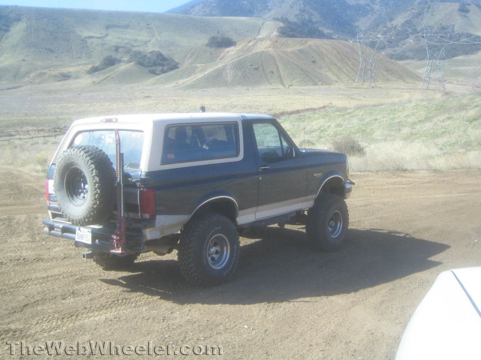 Dark Ford Bronco with white top and rear spare tire parked on a dirt road in the hills