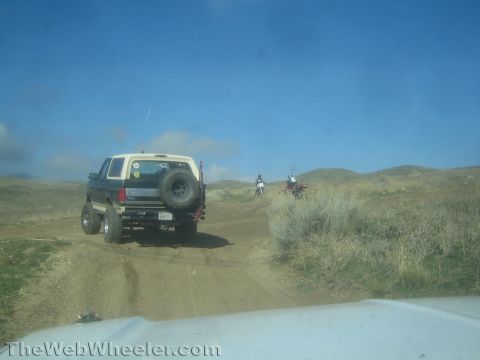 1990 Ford Bronco - lifted dark SUV with rear spare drives a dirt trail while two dirt-bike riders cross ahead across open scrubland under blue sky.
