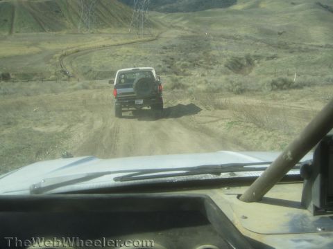 1990 Ford Bronco - Seen over a vehicle hood and wiper, a lifted SUV with rear spare tire drives a winding dirt trail through dry hills.