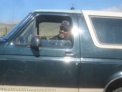 1990 Ford Bronco - smiling bearded man in a baseball cap sits in the driver's seat of a dark green Ford Bronco with white top, window down outdoors.