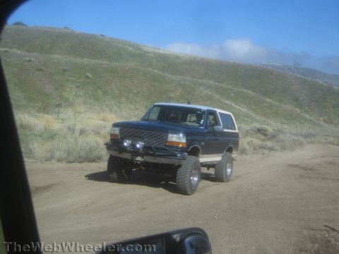 1990 Ford Bronco - lifted dark Ford Bronco with grille guard and round driving lights sits on a dirt road among dry hills, seen through a vehicle window.