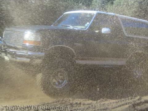 1990 Ford Bronco - lifted dark Ford Bronco with billet grille blasts through a muddy puddle, throwing up a heavy spray of brown water and droplets.