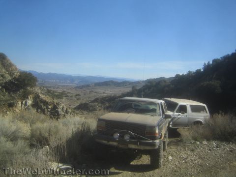 1990 Ford Bronco - Two lifted Ford Broncos, one dark and one tan, park on a high ridge dirt trail overlooking a hazy mountain valley below.