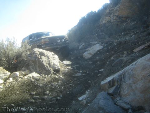 1990 Ford Bronco - dark SUV sits atop a steep, rocky off-road climb framed by boulders and brush against a bright sky in a high-desert canyon.