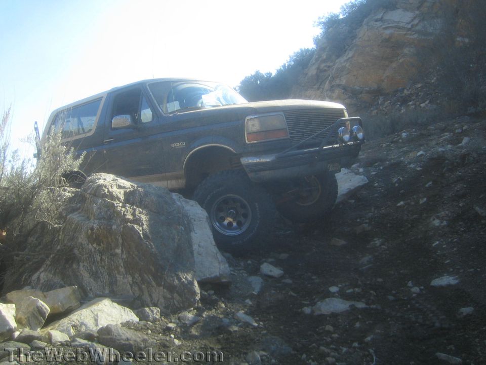 Lifted brown-and-tan Ford Bronco articulating over rocks on a hillside trail