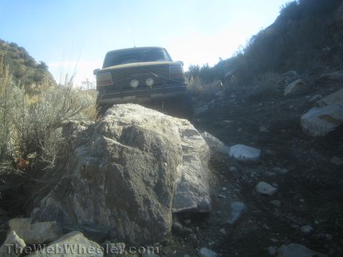 1990 Ford Bronco - Low front view of a lifted Ford Bronco with round driving lights climbing over a large boulder on a rocky high-desert trail.