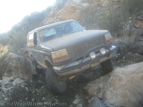 1990 Ford Bronco - dust-covered lifted Ford Bronco with chrome billet grille and round driving lights crawls a rocky trail flexing its front suspension.
