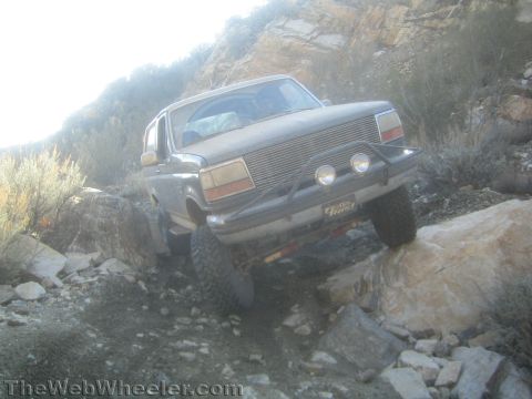 1990 Ford Bronco - lifted Ford Bronco with billet grille and driving lights climbs a steep rocky trail, front wheel lifted as it articulates over rocks.