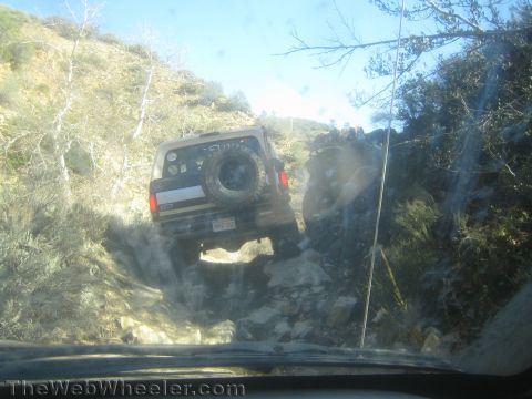 1990 Ford Bronco - Seen through a dirty windshield, a lifted SUV with rear spare tire climbs a rocky ledge on a brush-lined hillside trail.