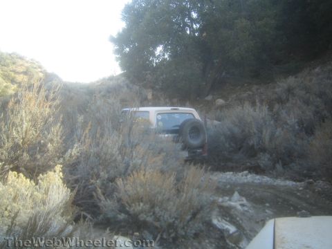 1990 Ford Bronco - backlit SUV with white top and rear spare tire works down a rocky, sagebrush-lined trail in a hazy sunlit canyon.